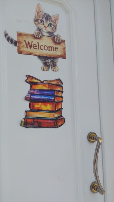 A decorative sticker of a kitten holding a "Welcome" sign and a stack of colorful books is affixed to a white door. The kitten is depicted with tabby markings, large eyes, and is positioned above the stack of books. The books are arranged in a slightly offset, vertical pile, showcasing spines in shades of red, orange, yellow, blue, and purple. The door has a visible handle on the right side, which is metallic and curved. The scene appears to be on the exterior of a building. The provided contextual information states this image is from Νέα Μηχανιώνα, Greece.