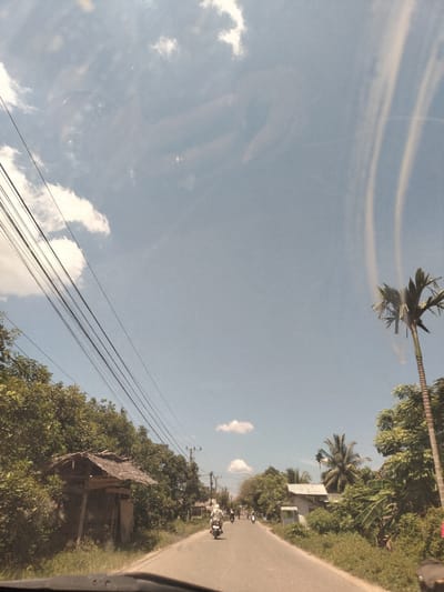 The image depicts a road in Meunasahbujok, Indonesia. The road is paved and straight, leading into the distance. There are people riding motorcycles on the road. The sky is blue with some clouds. There are trees and vegetation along the sides of the road, including a palm tree on the right. There is a small structure with a thatched roof on the left side of the road. Power lines are visible in the upper left portion of the image.