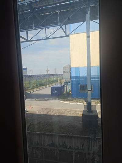 The image depicts an industrial area, possibly a port or manufacturing facility, viewed through a window. In the foreground, a concrete wall with barbed wire runs horizontally across the bottom of the frame. To the left and right, the dark frames of the window are visible.
Beyond the wall, a paved area is present, with a small orange traffic cone visible in the middle ground. A blue shipping container with Chinese text is located on the paved surface. In the background, large industrial buildings are present, one with a yellow and blue facade, and another with a grey metal structure. Overhead, a complex network of industrial scaffolding and elevated conveyor belts spans across the upper portion of the image. Electrical pylons are visible in the far distance. The sky is bright and appears to be partly cloudy. The scene suggests an active industrial zone. The location is identified as Дүңгілағаш, Kazakhstan.