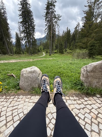 The image shows a pair of legs and feet wearing hiking boots and black leggings, positioned on a paved stone pathway. To the side of the pathway, there's a grassy area with two large rocks. Beyond the grass, there's a path leading into a forest of tall, evergreen trees under an overcast sky. This photo was taken in Brzegi, Poland, near the mountains.