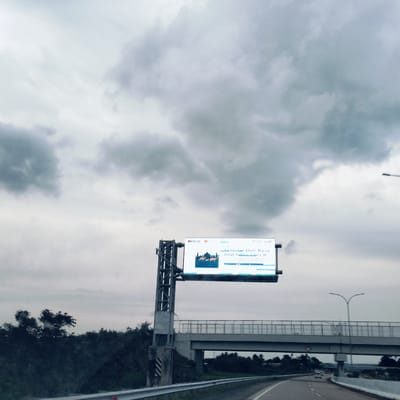 The image captures a highway scene under a heavily overcast, cloudy sky. The perspective suggests it was taken from a vehicle moving along the road.
In the foreground to the left, a tall metal structure supports a large electronic billboard. The billboard displays a message related to a religious holiday. The visible text reads "Selamat Hari Raya Idul Adha 144x H" (where '144x' likely represents a Hijri year, such as 1444 H). Below this text, there is an illustration featuring a silhouette of a mosque or eid ground with three animals, likely cattle, which are traditionally sacrificed during Eid al-Adha. Logos for "BANK JATIM" and another financial institution (possibly "BPR") are visible in the upper left corner of the screen.
An concrete overpass or bridge spans across the highway, supported by pillars, with guardrails running along its sides. Beneath and around the overpass, lush green foliage and trees are visible, extending into the distance on both sides of the road. A metal guardrail runs alongside the highway in the foreground. In the mid-ground, further down the highway, a white car can be seen. Streetlights with double arms are also present along the roadside.
The weather appears gloomy and muted due to the dense cloud cover. The time of day is daytime, though the lack of direct sunlight suggests it could be an early morning or late afternoon, or simply a very cloudy day. The setting is a road or tollway in Sihomlhok, Indonesia, indicated by the holiday greeting and the general infrastructure.