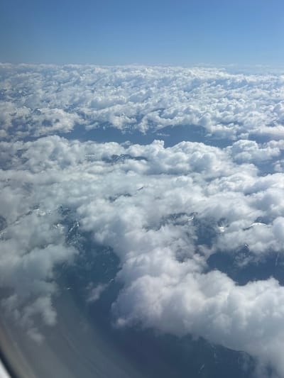The image presents an aerial view of clouds, seemingly taken from an airplane window. The left side of the image shows a clear blue sky transitioning into a dense layer of white, puffy clouds. Below the clouds, hints of snow-capped mountains can be discerned, suggesting a high-altitude perspective. There is no evidence or indication in this image of anything in Kelowna, Canada.