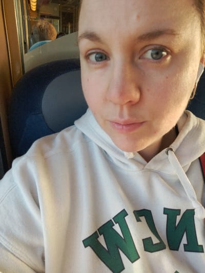 A female individual is depicted in a close-up, chest-up shot, presumably a selfie, inside what appears to be a train carriage in Fegersheim, France. She possesses a fair complexion, light blue-green eyes, and light brown or blonde hair pulled back. Her expression is neutral as she looks directly at the camera. She is wearing a white hooded sweatshirt with partial green block letters visible on the front, reading "NEW". A white wireless earbud is inserted in her right ear, accompanied by a small gold-colored hoop earring. To her left, part of a dark blue and grey upholstered seat is visible. In the reflection of the window behind her, the back of another passenger with short, light-colored hair, wearing a light blue top, is discernable, along with interior elements of the train carriage.