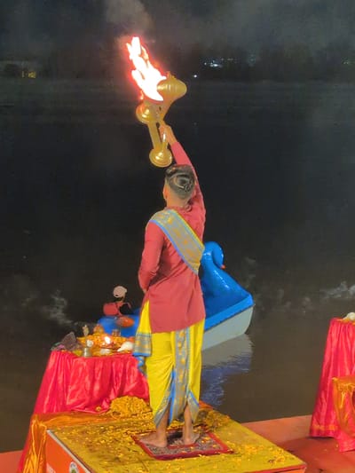 A rear view of a person in traditional Indian attire, including a maroon shirt and a yellow dhoti with blue trim, stands on a platform covered in yellow petals. The person holds a golden ceremonial lamp with flames aloft. The platform is situated near a body of water, possibly the Tawi River in Jammu, India, where a blue boat with a decorative swan head is visible. In the background, lights twinkle, suggesting the scene occurs at night or dusk. To the left, a red table is draped with a red cloth and adorned with flowers and religious items, including a conch shell and a lit oil lamp. A red cloth-covered table is also visible on the right. The overall atmosphere suggests a religious ceremony or ritual.