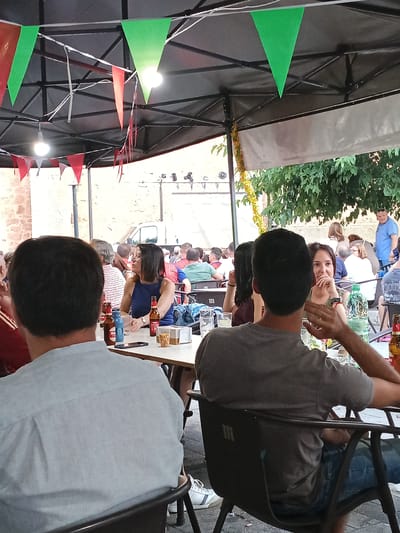 The image depicts an outdoor gathering of people seated at tables under a tent structure. Red and green triangular flags are strung across the ceiling of the tent. In the foreground, two individuals are visible from behind, seated at a table. The person on the left wears a light blue collared shirt, and the person on the right wears a grey t-shirt and blue jeans. On the table between them are several bottles of beer, a blue cup, a mobile phone, and a clear plastic cup containing snacks. A woman in a blue dress with glasses is seated at a table to the right, looking towards the camera. In the background, other patrons are seated at tables, and a white van is visible. The setting appears to be an outdoor dining or drinking area, likely in a Spanish town, possibly Campillo de Altobuey, given the context. The overall atmosphere suggests a casual social event.