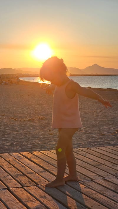 A child, seen from the side and from the waist down, stands on a wooden walkway at the beach. The child is wearing a pink sleeveless shirt and shorts, and is looking down, with their arms outstretched to the sides as if balancing. The sun is setting, casting a warm orange glow over the scene. The ocean is visible in the background, with a faint outline of mountains on the horizon. The beach is sandy, with a few people and some beach umbrellas in the distance. The wooden walkway is made of planks laid diagonally.