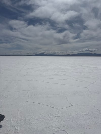 The scene depicts an expansive view of the Salar de Uyuni in the Municipio Colcha K, Bolivia. The foreground and midground consist of a vast, uniformly white salt flat, characterized by distinctive polygonal cracking patterns across its surface. Faint, linear impressions consistent with vehicle tracks are visible on the salt. The horizon line is flat and distant, where the salt flat meets a low, dark mountain range stretching across the entire width of the background. The sky above is partially overcast with a mix of grey and white clouds, interspersed with patches of clear blue. A small, dark, unidentifiable object, possibly part of footwear, is present in the extreme lower-left corner of the frame. No individuals, specific actions, or interactions are depicted. The environment is open and desolate under natural daylight.