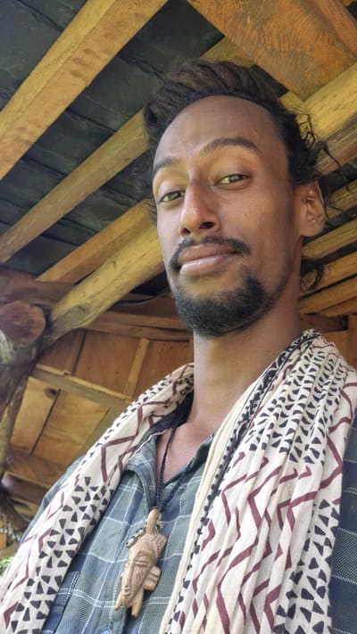 A close-up, head-and-shoulders selfie shows a man with dark curly hair and a goatee, looking towards the camera. He has light green eyes and is wearing a plaid shirt, a patterned scarf, and a wooden pendant necklace. The background consists of a wooden roof structure with exposed beams and dark roofing material, typical of outdoor shelters found in rural areas of Shila, India. The lighting suggests an outdoor setting during the daytime.