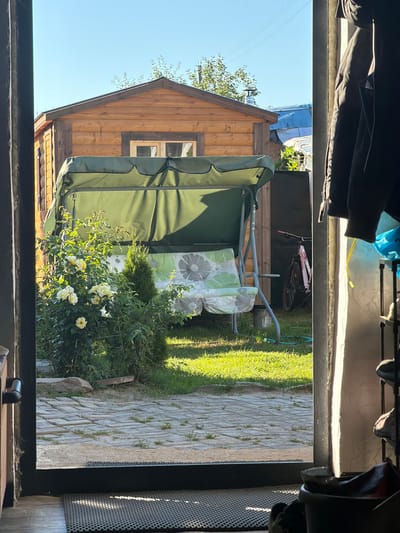 The image depicts a view from an interior doorway looking out onto a sunny backyard. In the foreground, a dark doorway frame is visible, with a black doormat on the floor just outside. The backyard features a small wooden shed with a gable roof and a window. In front of the shed is a green garden swing with a floral-patterned cushion. A bush with white roses and a conifer are to the left of the swing. The ground is covered with cobblestones and patches of green grass. A bicycle is leaning against the shed in the background, and further back, there are other structures with blue and white roofs. To the right of the doorway, several jackets are hanging, and a shelf with shoes is visible. The scene is set in a rural environment, likely in the Валевачскі сельскі Савет region of Belarus.