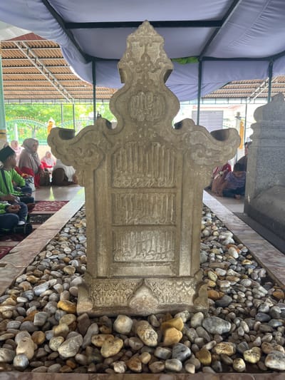 The image depicts a tombstone in the city of Beuringen, Indonesia. The tombstone, carved from light-colored stone, is intricately designed with Arabic calligraphy. It is surrounded by a bed of smooth, rounded pebbles of various colors, including grey, white, and yellow. The tombstone is situated outdoors, beneath a canopy structure supported by metal beams and covered with a light purple fabric. In the background, several individuals, including men, women, and children, are seated on the floor, some wearing traditional Islamic attire. The ground appears to be tiled, with a red carpet visible in the foreground. The overall environment suggests a sacred or commemorative site, possibly a cemetery or a shrine.