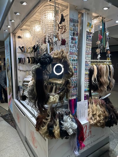 A beauty kiosk showcases hair extensions in various colors and styles. Wigs with shades of blonde, brown, and black are neatly arranged alongside colorful hair clip-ins. Shelves below display sparkling hair accessories and jewelry. A ring light and illuminated pendant lights contribute to the bright ambiance.