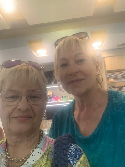 Two women pose for a selfie in what appears to be a commercial establishment with shelves stocked with products. The woman on the left wears glasses, a patterned shirt, and a gold chain. The woman on the right has blonde hair and wears a teal shirt and hoop earrings. The lighting in the image is bright, with some glare. The background suggests a retail environment, possibly a small store or convenience shop. The location in Odivelas, Portugal is not discernible from the image itself.