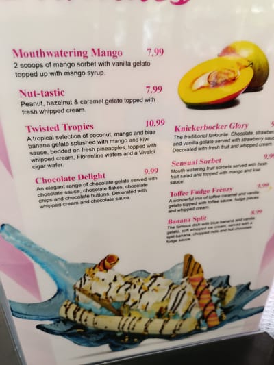 This is an image of a menu featuring various ice cream and gelato desserts. The menu items are described with their ingredients and prices.
Visible text includes:
- "Mouthwatering Mango" with a price of "7.99"
- "Nut-tastic" with a price of "7.99"
- "Twisted Tropics" with a price of "10.99"
- "Chocolate Delight" with a price of "9.99"
- "Knickerbocker Glory" with a price of "9.99"
- "Sensual Sorbet" with a price of "9.99"
- "Toffee Fudge Frenzy" with a price of "9.99"
- "Banana Split" with a price of "8.99"
There are also illustrative images of some of the desserts. One image shows a halved mango with another whole mango behind it. Another image displays a colorful ice cream dessert with whipped cream, chocolate drizzle, and wafer cookies.
The context indicates the location is Bedford, United Kingdom. The image appears to be a photograph of a physical menu, possibly in a cafe or restaurant. There are no people or specific time of day or weather cues visible. The overall tone is appetizing due to the descriptions and images of desserts.