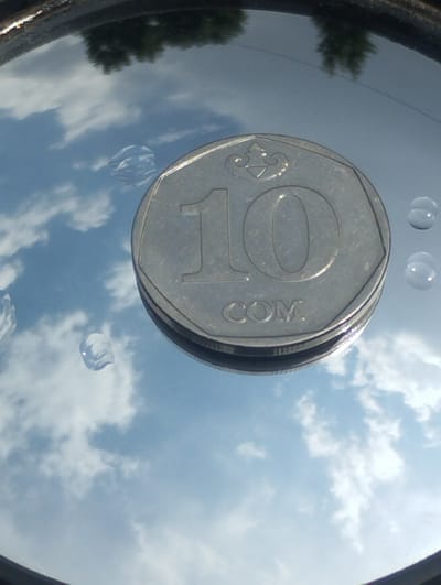 The image shows a close-up, angled view of a ten-unit coin, possibly a Russian Ruble or a similar denomination, lying on a reflective surface. The coin is silver-colored and shows the numeral "10" prominently on its face, along with some stylized design above it and the letters "COM" below. The reflective surface shows a clear reflection of a blue sky with scattered white clouds, and the dark silhouettes of trees are visible at the top edge of the image. There are also a few water droplets on the reflective surface, which appear to be around the coin. The overall scene suggests an outdoor setting, possibly in Vinogradnoe, Kyrgyzstan, where the coin is placed on a surface like glass or polished metal, reflecting the surrounding environment. The image is shot with a slightly low angle, emphasizing the coin and the reflected sky.