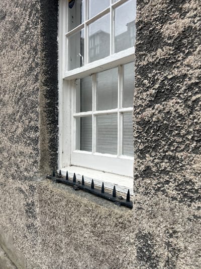 Captured in Aberdeen City, United Kingdom, the image presents a close-up view of a window set within a textured building facade. The window, painted white, features a grid pattern of panes and appears slightly aged, with minor signs of wear such as small cracks. To the right of the window, a black metal deterrent device with protruding spikes is affixed to the building's exterior, presumably to discourage birds or other animals. The building's facade exhibits a rough, speckled texture in varying shades of gray, contributing to an overall impression of age and weathering. The composition suggests a static scene focused on architectural details.