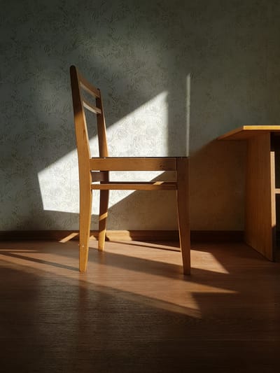 The image depicts an interior scene, likely a room, with a wooden chair positioned prominently in the foreground. The chair is made of light-colored wood and has a simple design with a slatted back and seat. Sunlight streams in from an unseen source, casting a sharp diagonal shadow across the chair and the patterned wallpapered wall behind it. The light also illuminates the wooden floor, creating a warm glow and distinct shadows from the chair's legs. To the right of the chair, a portion of a wooden table is visible. The wallpaper has a repeating, intricate floral or organic pattern. The overall impression is one of quiet solitude and the play of natural light in an ordinary setting. There are no people present in the image, and no actions or interactions are occurring. The scene appears to be a snapshot of an empty room, with the focus on the light and shadows. The image does not provide any specific visual cues to indicate its location in the city of Kurchatov, Kazakhstan.