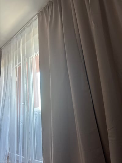 A close-up, low-angle shot shows a portion of a window with two layers of curtains. The left side features a sheer white curtain, allowing light to filter through to the windowpane and the outside. To the right, a thicker, opaque curtain hangs, obscuring the majority of the window and casting shadows. The curtains are suspended from a white ceiling track. The scene is illuminated by natural light coming from the window. The image does not depict any people, actions, or interactions. The location is identified as Gebersdorf, Austria, although no specific landmarks or contextual cues within the image confirm this.