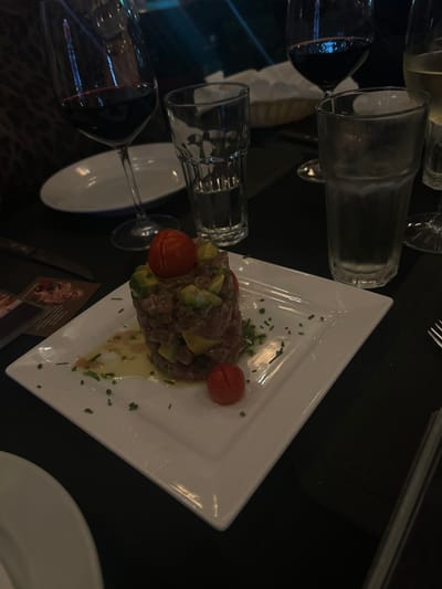 A white square plate sits on a black tablecloth, in a dimly lit restaurant setting, with the coordinates 9.935, -84.189, placing it in Santa Ana, Costa Rica. The plate contains a layered appetizer, likely tuna tartare, adorned with diced avocado, chopped chives, and two cherry tomatoes. A glass of red wine and a glass of water are visible in the background, along with a half-empty glass of white wine out of focus. The table has a dark brown tablecloth, with two white plates near the edge of the frame. A fork is partially visible in the lower right corner. The restaurant atmosphere is intimate and casual.