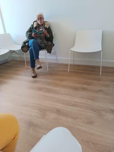 An older woman with short white hair and glasses is seated in a brightly lit modern waiting room, engrossed in reading a small book. She is wearing a dark green jacket with a cream-colored faux fur collar, a plaid scarf, blue jeans, and tan ankle boots. Her legs are crossed at the ankles. She sits on a white, minimalist chair with thin metal legs, positioned against a plain white wall.
To her right, an identical empty white chair stands. Another white chair is partially visible to her left. The room features light wood-patterned flooring, likely laminate or vinyl, with white baseboards along the walls. Natural daylight illuminates the scene, suggesting it is daytime. In the foreground, parts of a yellow chair and another white chair are visible, indicating the perspective is from someone also seated in the room. The book she is reading has a turquoise or light blue cover, with the word "VIVRE" partially legible on it. The setting appears to be a public waiting area in Thionville, France. The woman appears calm and focused on her reading.