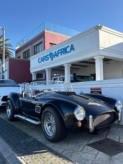 A black classic sports car, likely a Cobra, is parked on a paved surface in front of a building with "CARS4AFRICA" signage. The building features white columns and a burgundy and beige facade. A white SUV is visible behind the sports car, and a palm tree is to the left. A "WARNING" sign is attached to a white wall in the foreground. The scene is set in Blouberg, South Africa, under a clear blue sky.