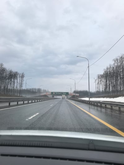 The image shows a view from inside a car driving on a wet highway. The road is straight and appears to be made of asphalt. There are white and yellow lines marking the lanes. Guardrails run along both sides of the road. Bare trees are visible on both sides of the highway, with some remaining snow near the guardrail on the right. An overhead sign reads "TATAPCTAH", a Russian spelling of "Tatarstan". This is a bridge with some type of sign. The sky is overcast, creating a gray, muted atmosphere. The location is likely near Staroe Tyaberdino, Russia, which is implied by the "Tatarstan" sign, as Tatarstan is a republic within Russia. The road appears to be wet from recent or ongoing rain. No people are visible in the image.