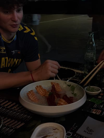 The image depicts a close-up, nighttime view of a person sitting at a table with a plate of sashimi. The person, on the left, is wearing a navy blue sports jersey with yellow accents and the brand name "hummel" is visible. They have short, curly brown hair and are looking towards the right of the frame. Their left arm is resting on the table, and a red string bracelet is visible on their wrist.

In the center of the frame is a white bowl filled with sashimi, served on a bed of crushed ice. The sashimi appears to be salmon and tuna. To the right of the sashimi bowl is a small white dish containing green wasabi. A green glass bottle of Coca-Cola is also visible on the right side of the table, along with chopsticks resting on the table. The table is covered with a dark tablecloth, on which a menu with various food items and prices is spread out. The background is dark and out of focus, suggesting an outdoor setting, possibly a restaurant patio.

While the image captures a dining scene, it does not contain specific visual cues to identify the location as Premià de Mar, Spain.