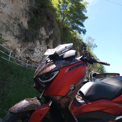 The image displays a red Yamaha XMAX scooter parked outdoors, possibly in Kalalengkio, Indonesia, given the context provided. The scooter is positioned on a grassy embankment next to a rocky cliff face with a metal guardrail. The scooter has a custom wrap with a red, black, and gold fragmented pattern. It features a black seat, LED headlights, and a license plate that reads "BL 5711 VNJ". The background shows lush green vegetation, trees, and a bright blue sky. The lighting suggests it is daytime, likely sunny. No people are visible in the frame.