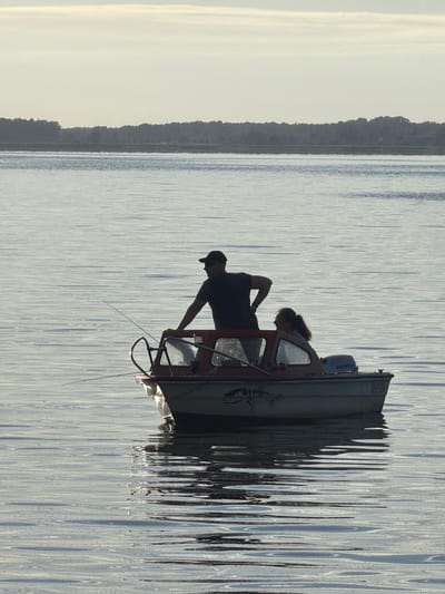 The image shows a side view of a small fishing boat on calm water. The boat is oriented vertically in the frame, suggesting the photo may have been rotated. Two people are visible in silhouette within the boat. One person, likely male, is wearing a baseball cap and appears to be leaning over something inside the boat. Another person, possibly female, with long hair pulled back, is standing beside them, looking down. The boat has a cabin with a windshield and a dark-colored interior. A Honda outboard motor is mounted on the back of the boat. The water is serene with subtle ripples, and the sky is a pale, bright color, indicating it could be early morning or late afternoon. The reflection of the boat and the surrounding environment on the water is distorted by the ripples, creating an abstract pattern. On the left side of the image, a dark, tree-lined shore is visible, suggesting a lakeside or coastal setting. The overall mood is peaceful and quiet. There is some visible text on the stern of the boat that appears to be a license plate number, reading "ABD-X531". The location context is Ribnitz-Damgarten, Germany.