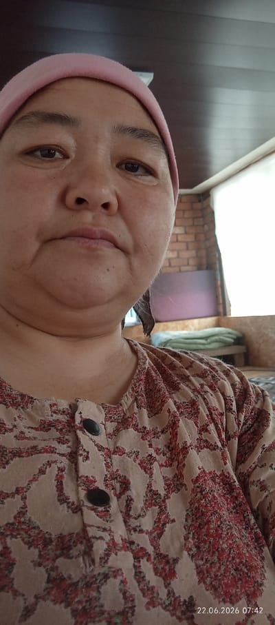 A head and shoulders selfie, taken indoors, shows a middle-aged woman wearing a pink headscarf and a light brown blouse with a red floral pattern. The woman is looking towards the camera with a neutral expression. The background appears to be a room with a wooden ceiling and a window with sheer white curtains. A brick wall is visible behind the window, and a bed with light green blankets is in the distance. The bottom right corner of the image shows a timestamp: "22.06.2026 07:42". The location is identified as Chon-Kara, Kyrgyzstan.