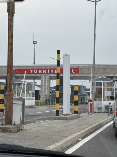 The image depicts the Turkish entry point of the Lesovo-Hamzabeyli border crossing, geographically associated with Lesovo, Bulgaria. The vantage point is from inside a vehicle positioned in a queue lane.
The scene is dominated by a grey overhead archway spanning multiple lanes. A prominent red band on the archway displays "TÜRKİYE" in white capital letters, flanked by two Turkish flag emblems (white star and crescent on red). The archway's supports feature a repeating grey geometric pattern.
Multiple black and yellow striped bollards delineate vehicle lanes. A tall, white, rectangular structure is centrally positioned in one lane, flanked by these bollards. To the left, a white-framed glass guard booth is visible with its white barrier arm in a raised position, and yellow metal railings are behind it. To the right, another white-framed glass guard booth, partially visible, has an external air conditioning unit. An orange traffic cone stands near this right booth.
In the immediate left foreground, a rusted brown metal pole rises vertically. A tall, grey metal street light pole is on the right side of the frame. In the background, a tall, grey lattice-style communication tower is visible above the archway.
The roadway is asphalt with white lane markings. A grey concrete slab sidewalk borders the right side of the road. A small white piece of litter is visible on the ground next to the sidewalk curb. The rear of a white sedan is partially visible on the far right. The sky is uniformly overcast and light grey. No people are discernible in the frame.