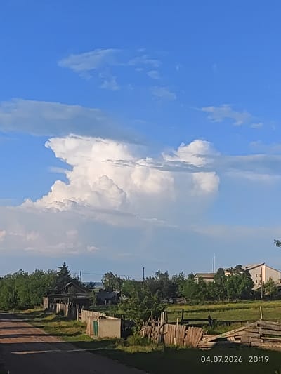 The image shows a rural landscape in Konovalovo, Russia, under a partly cloudy blue sky. In the foreground, a dirt road leads towards a cluster of weathered wooden structures and fences. Trees and foliage are abundant throughout the scene. In the background, a large, towering cumulus cloud dominates the sky. A date and time stamp "04.07.2026 20:19" is visible in the lower right corner of the image. There are no people visible in the photograph.