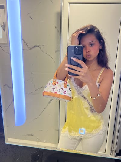 A young woman is taking a mirror selfie in what appears to be a bathroom with marble walls and a blue illuminated panel. She is wearing a yellow camisole with white lace trim and white pants. She is holding a black smartphone in her right hand and a small, multi-colored designer handbag in her left hand. She has long, dark hair and is wearing hoop earrings and a bracelet. The setting does not provide any information to determine the location in Chaidefto, Greece.
