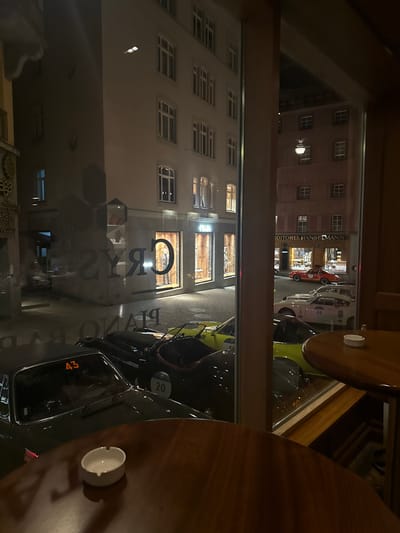 The scene is viewed at night from inside the Grys Piano Bar in Sankt Moritz, Switzerland. In the immediate foreground, a dark wooden table with an empty white ashtray is visible. Beyond a large window, a paved street is occupied by multiple classic sports cars. These include a dark green car with '43' displayed on its windshield, another dark vehicle with '20' on its side, a lime green car, a white rally car marked '24', and a red vintage sports car. Reflections on the window show parts of the 'GRYS PIANO BAR' signage and a 'PRADA' storefront on the left. Directly opposite, a multi-story building with a reddish-brown textured facade features numerous windows and a brightly illuminated sign for 'CONDITOREI HANSELMANN' above its entrance. No people are visible.