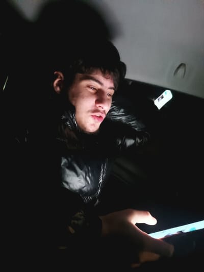 A young man with dark hair and fair skin looks at a glowing phone in his hands. He wears a black, shiny puffer jacket and seems to be sitting in a vehicle at night. An overhead light reflects on the car's interior. The man's face is partially illuminated by the phone's light, casting shadows across his features. The scene is dark, with the man and phone being the primary source of illumination.