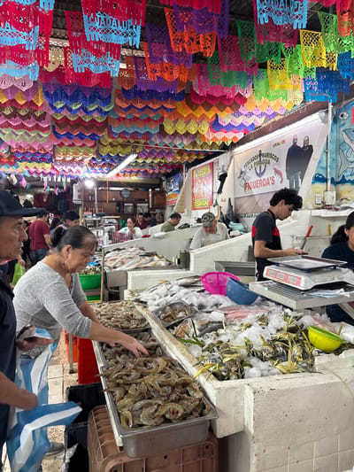 A bustling indoor market scene. Overhead, colorful paper banners and decorations fill the space. Customers browse a seafood stall piled high with ice, fish, crabs, and shrimp. Vendors stand behind the counter, one using a scale. The market appears crowded with people further down the aisle.