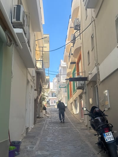 The image depicts a narrow, stone-paved alleyway in Agios Nikolaos Municipal Unit, Greece. The buildings lining the alley are whitewashed, with several balconies and windows visible. The time of day appears to be daytime, with bright sunlight casting shadows.

The main subjects are a person walking away from the camera down the alley and a parked black motorcycle. The person is wearing dark clothing. The motorcycle has a license plate that reads "AEE 709 GR". Other objects in the scene include air conditioning units mounted on the buildings and electrical wiring running along the walls. There is also some signage visible on a building in the distance.

The overall atmosphere is that of a quiet, sun-drenched Mediterranean street. The scene is tranquil, with no other people or activity immediately apparent, suggesting a peaceful moment in the day. The stone paving and architecture are characteristic of a Greek island or coastal town. The clear blue sky suggests good weather.