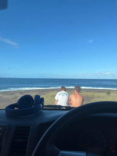 A partial view from inside a vehicle shows a beach scene. Two people stand near the water; one is shirtless and the other wears a white t-shirt. The ocean extends to the horizon under a clear blue sky. The vehicle's dashboard, including the steering wheel, speedometer, and a pair of blue headphones are visible, framing the outdoor scene.