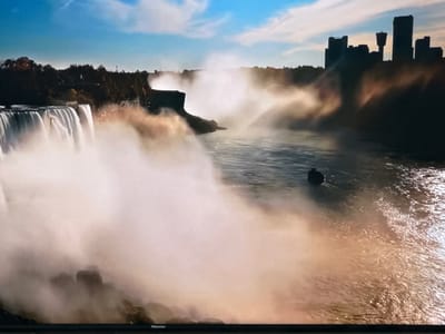 The image depicts a wide, powerful waterfall cascading into a turbulent river. Mist rises from the impact of the water, creating a hazy atmosphere. In the distance, silhouetted against a bright sky with scattered clouds, is a city skyline characterized by tall buildings. The river's surface is disturbed by the flow and reflects the warm tones of the mist. The bottom of the image shows the edge of a television screen with the brand "Hisense" visible. The location described as Ləcət, Azerbaijan, is not identifiable in the image itself.