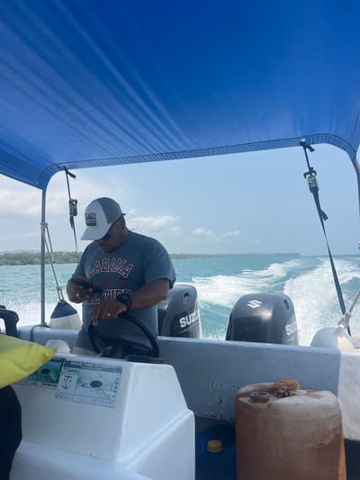 A medium, eye-level shot shows a man driving a boat at sea. The man is wearing a grey t-shirt with "ALABAMA" printed in red and white, a grey baseball cap, and a watch. He appears to be looking at something in his hands while steering the boat. Behind him are two Suzuki outboard motors. To the right of the motors is a large, tan container.

The boat is moving quickly, as evidenced by the wake in the water. The sky is blue with some clouds. A blue canopy covers part of the boat. The scene appears to be taking place near the coast of Livingston, Guatemala, as a sticker that reads "BELIZE PORT AUTHORITY BELIZE BOAT LICENSE A46151" on the boat’s dashboard suggests the location.