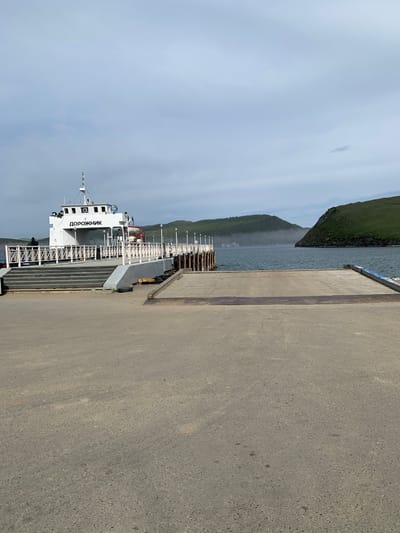 In Sakhiurta, Russia, on a cloudy day, a white ferry named "ДОРОЖНИК" is docked at a pier. The ferry has a multi-level superstructure with windows and is equipped with safety railings. A ramp extends from the pier to the ferry's deck, and stairs lead up to the ferry's passenger area. The pier is constructed of concrete and wood, with wooden pilings visible in the water. Beyond the pier, a body of water is visible, with green, hilly islands in the distance. The sky is overcast with a few wispy clouds. The overall scene depicts a typical ferry terminal setting.