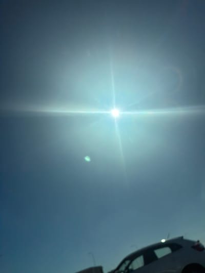 The image captures a bright, sun-drenched sky with a partial view of a white vehicle. The sun is positioned in the upper-left quadrant, appearing as an intense, brilliant white orb with strong lens flare extending horizontally and vertically across the frame, creating a distinct starburst effect. A faint, circular halo artifact is visible above and slightly to the left of the sun, and a smaller, greener lens flare spot is present to the right. The sky transitions from a very bright, almost blown-out white around the sun to a hazy blue-gray further away from the light source.
In the upper-right corner, a portion of a white vehicle is visible, viewed from an unusual low angle. It appears to be the rear section of a car or similar automobile, possibly showing a white bumper, a dark-colored tire, and potentially a taillight or reflector near the top edge. The orientation of the vehicle suggests it might be positioned on a lift in a workshop, or perhaps inverted.
The scene takes place during bright daytime, likely mid-day, given the high intensity and position of the sun. The weather appears clear and sunny, although the direct sun creates significant atmospheric haze or lens effects. No specific activity beyond the act of photographing the sun and the vehicle is explicitly depicted. There is no visible text within the image, and no specific location cues other than the implied location of Goodwood, South Africa.