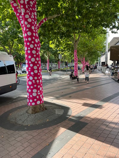 The image displays a vibrant, sunlit pedestrian promenade in Southbank, Australia. A prominent feature is a long row of trees lining the path, with their trunks and lower branches strikingly wrapped in bright pink fabric adorned with large white polka dots. This distinctive decoration creates an artistic and whimsical ambiance along the walkway, extending from the foreground into the midground.
The pathway is paved with light reddish-brown rectangular tiles, intersected by darker grey strips that run parallel to the tree line. Strong, defined shadows are cast across the pavement, indicating bright, sunny weather. The trees are full of lush green foliage, providing natural canopy over the area.
Several people are visible in the scene. In the midground, an individual walks along the path. Further down, two people are seated at a small outdoor table, suggesting a cafe or public seating area where visitors can relax. Other less distinct figures are visible in the background. A white van is partially visible in the bottom-left corner, parked near one of the decorated trees. Bicycles are also parked near some of the tree trunks along the promenade. The overall impression is one of a lively urban space, enhanced by the unique public art installation on the trees, under clear daylight conditions. No visible text is clearly legible.