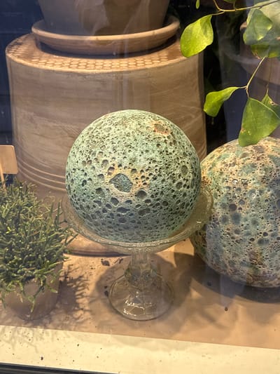 The image depicts a still life display, likely in a shop window, featuring decorative spheres and plants. The central focus is a large, textured sphere with a mottled green and brown surface, resting on a clear glass pedestal. To its right is another similar, but slightly larger, sphere, partially visible. In the foreground to the left, a small potted plant with thin, green branches is situated. In the background, several large, beige ceramic pots are visible, one with a ribbed texture and another holding a plant with broad green leaves. The overall scene is viewed through a glass pane, indicated by reflections and smudges on the surface. The location is identified as Frederiksberg, Denmark.