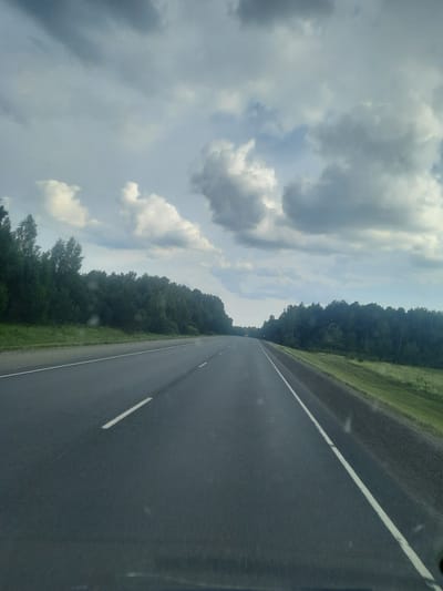 This media file captures a view from a moving vehicle on a paved road in Petrovka, Russia. The perspective is from the passenger seat, looking towards the right side of the road. The road itself is dark asphalt with a white dashed line on the right edge, indicating lanes.

To the left of the road, a dense, dark green forest stretches as far as the eye can see. The trees appear tall and closely packed, forming a solid wall of foliage. Beyond the forest, the sky is filled with dramatic, grey and white clouds. These clouds suggest a dynamic weather system, possibly a partly cloudy day with a chance of rain, or the aftermath of a recent shower. The lighting indicates it is daytime, likely afternoon, given the intensity of the light and the shadows cast by the clouds.

The overall mood is one of travel and passage through a natural, somewhat wild landscape. The composition, with the road leading into the distance and the expansive sky, evokes a sense of journey. There are no people or distinct objects visible besides the road and the natural elements of forest and sky. No text is visible within the frame.