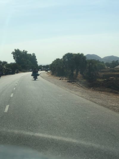 The scene depicts an outdoor environment on a paved road in Bundot, Nigeria, under a clear, bright sky. A dark-colored motorcycle, with at least two individuals riding, is positioned on the asphalt road, moving away from the viewer. The road displays a dashed white line on its left side and curves towards the left in the distance. To the right of the road, a gravel and dirt shoulder is present, with several piles of dark earth visible beyond it. A line of distinct palm-like trees runs parallel to the right side of the road. In the background, beyond these trees, faint structures and a range of hills are discernible. Denser green foliage lines the left side of the roadway. Other vehicles are faintly visible in the far distance along the road's curvature.