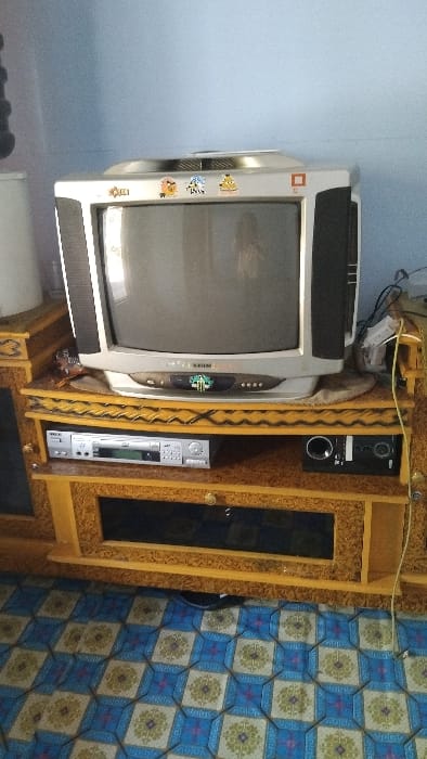The image shows a television set on a wooden entertainment center in a room with blue and white patterned linoleum flooring. The television, an older model, has stickers on the top. There is a DVD player and a black speaker underneath. The walls are a pale blue. The coordinates provided place the location in a residential area of the Kecamatan Panjalu district, Indonesia.