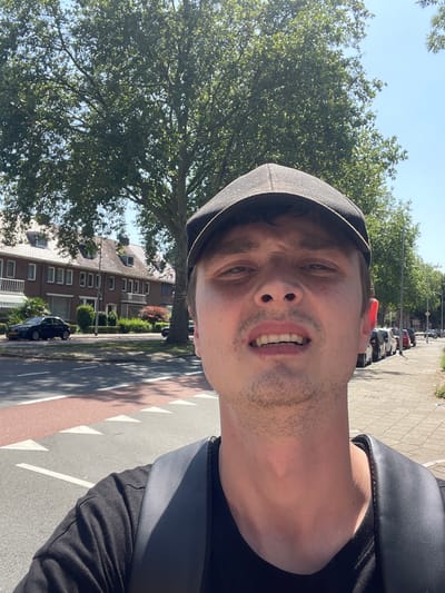 The media file is a selfie taken by a person wearing a black cap, black t-shirt, and a black backpack. The person is looking directly at the camera with a surprised or shocked expression. The image is taken outdoors during the day, with a clear blue sky visible. The background shows a tree-lined street with houses and parked cars. The sun is casting shadows, indicating it is daytime. The scene appears to be a residential area, possibly in Venlo, Netherlands, given the architecture of the houses and the overall environment. The lighting suggests it is a sunny day. The focus is on the person's face and upper body, with the background slightly blurred. No visible text is present in the image.