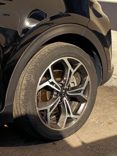 The image shows the rear quarter panel and wheel of a black Kia vehicle, likely in an outdoor setting. The car's paint exhibits some damage, with a noticeable dent on the body panel above the wheel arch. The wheel is a multi-spoke alloy rim with a silver and black finish. The tire appears to be a Bridgestone. The background consists of a textured wall and a patch of asphalt or gravel on the ground. The lighting suggests it is daytime, with shadows cast on the ground, indicating sunlight. There are no people or visible actions occurring in the image. The location is identified as Tash-Dobo, Kyrgyzstan.