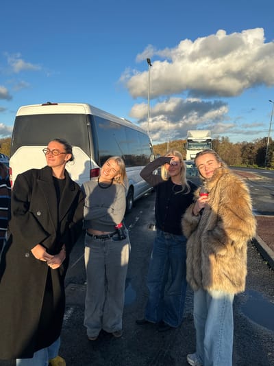 An outdoor scene in South Ockendon, United Kingdom, depicts four females standing on an asphalt surface under a clear blue sky with scattered white clouds. The sun is low, casting long shadows and causing some individuals to squint.

From left to right:
1.  A female, appearing to be in her 20s-30s, stands wearing a dark, knee-length double-breasted coat over a dark top and light-colored trousers. She wears eyeglasses and has dark hair tied back. Her eyes are closed or squinting, and her hands are clasped at waist level.
2.  A female, appearing to be in her 20s, stands with arms crossed, wearing a grey long-sleeved top, grey wide-leg jeans, and a dark beaded necklace. She is smiling broadly. A red Coca-Cola bottle is partially visible, tucked into her right front pocket.
3.  A female, appearing to be in her 20s, wears a dark cardigan over a dark top and blue wide-leg jeans. Her right hand is raised, shielding her eyes from the sun.
4.  A female, appearing to be in her 20s, wears a long, light brown faux fur coat over light blue wide-leg jeans. She holds a red cup in her left hand, and her eyes are closed or squinting.

In the background, a white mini-coach/bus is parked behind the individuals. Further back on the right, a white semi-truck/lorry cab is visible. Multiple dark-colored lampposts are present. The ground is an asphalt lot with some visible puddles of water. Distant dark vegetation lines the horizon.