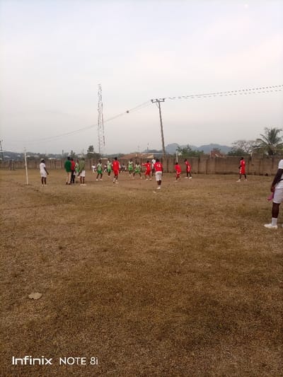 A soccer game is in progress on a dry, grassy field. Players in red jerseys and green jerseys are actively engaged. A ball is airborne in the center of the field. In the background, an electrical tower and utility poles are visible, with foliage and a hillside adding depth. The sky is overcast. A goalpost marks the left side of the frame. The lower part of the image also has "Infinix NOTE 8i" written in bold white letters on it.